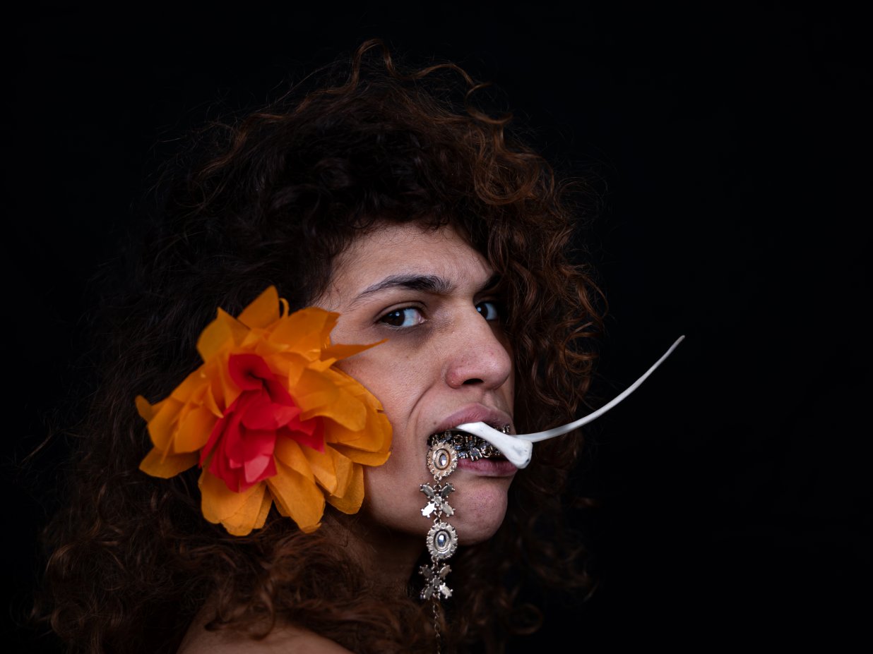 eine Frau schaut seitlich in die Kamera, sie hat eine gro&szlig;e tropische Bl&uuml;te im Haar, aus ihrem Mund quillen Goldschmuck und Tierknochen