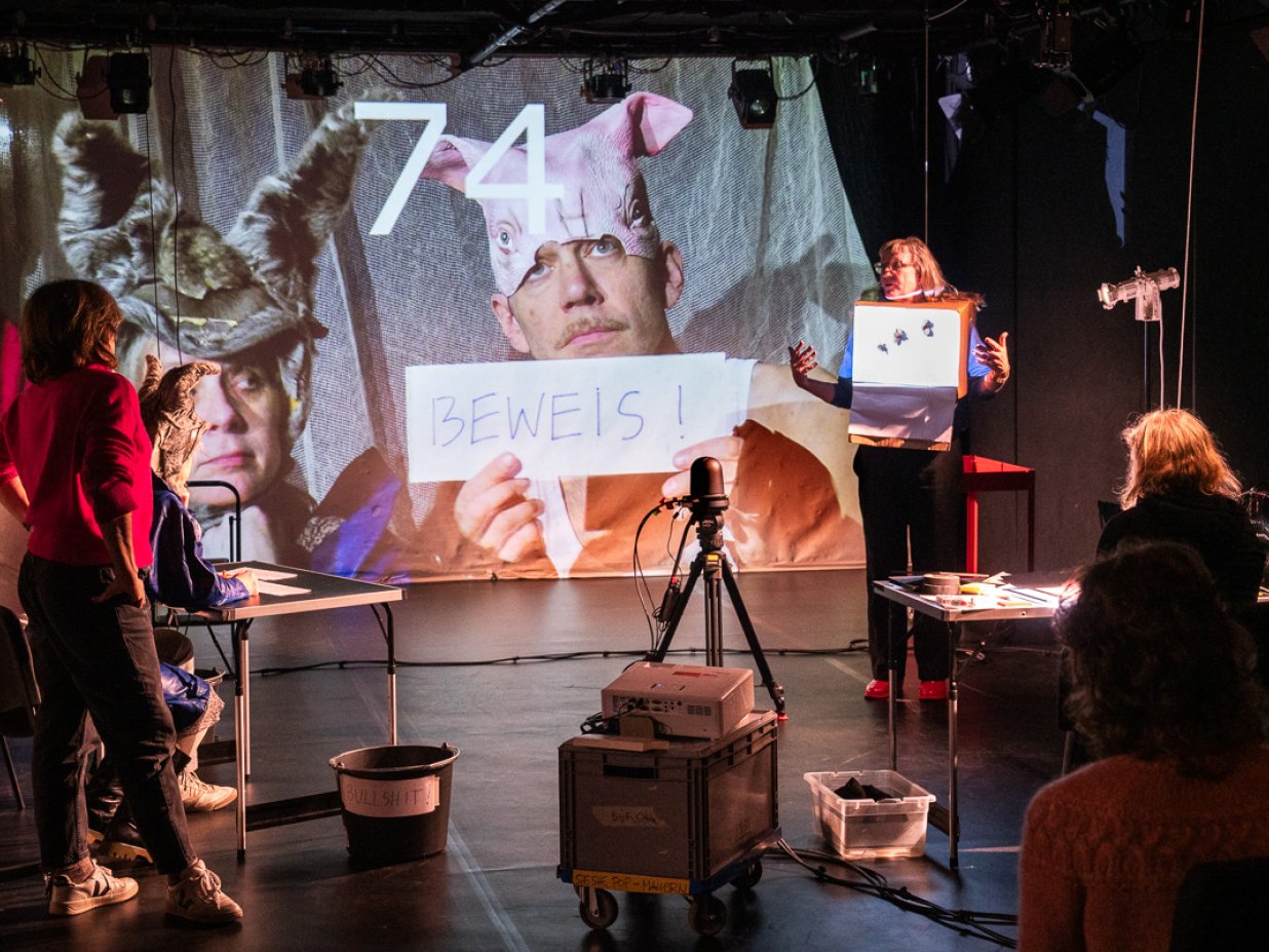 Im B&uuml;hnenraum steht eine Anordnung von Stativen und Kisten.  Mehrere Performerinnen schauen einer Performerin zu wie bei einem Vortrag. An der B&uuml;hnenr&uuml;ckwand ist ein Kamera Bild von 2 Person