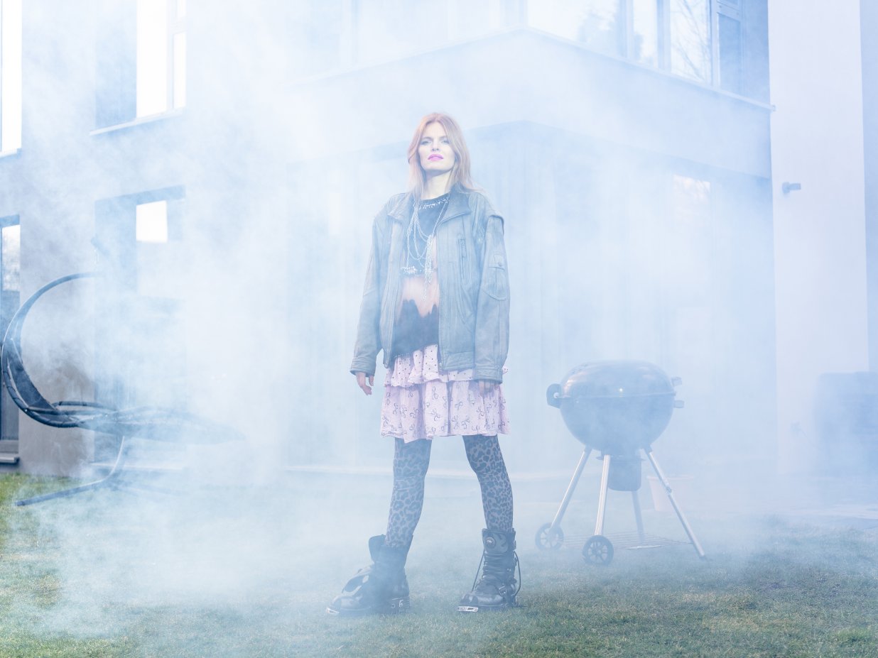 Eine punkig gekleidete Frau steht im Nebel vor einem Einfamilienhaus mit Grill.